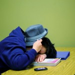 student sleeping hunched over desk with hat on their head
